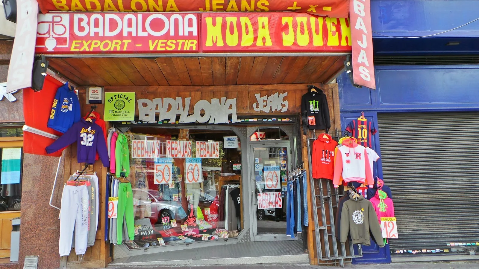 Fachada real de Vaqueros Badalona en la Ctra. de Canillas: rótulo rojo y amarillo «BADALONA · MODA JOVEN», letras blancas en relieve «BADALONA JEANS», y el escaparate de rebajas con vaqueros, sudaderas, chándales y camisetas colgados.